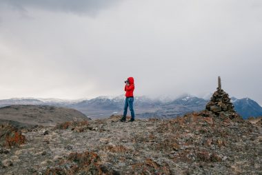 man in red jacket standing using telescope while standing on a cliff of a mountain